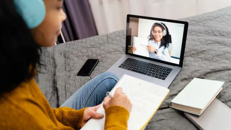 Mulher com fones de ouvido participa de uma videochamada no notebook, assistindo uma professora que mostra material didático.