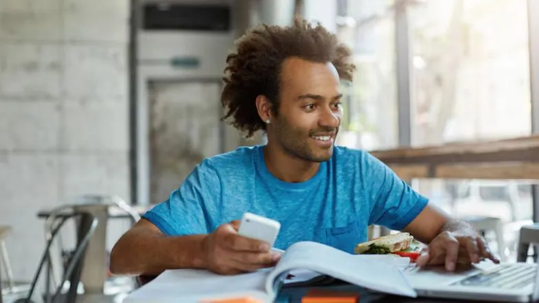 Jovem sorridente usando celular e laptop enquanto estuda com livros, transmitindo flexibilidade no processo de ingresso e estudo.
