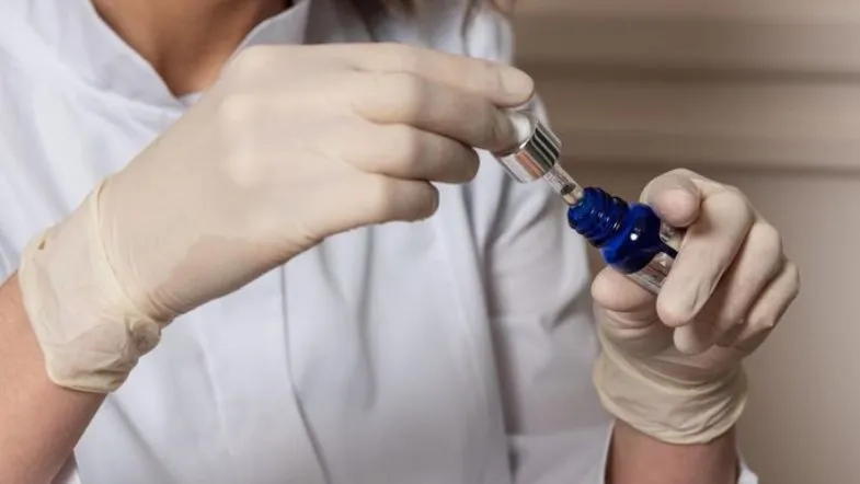 O que é Biomedicina Estética? Formação, regulamentação e áreas de atuação 12 Profissional com luvas manipulando frasco de dermocosmético azul com conta-gotas, simbolizando uso de ativos na biomedicina estética.