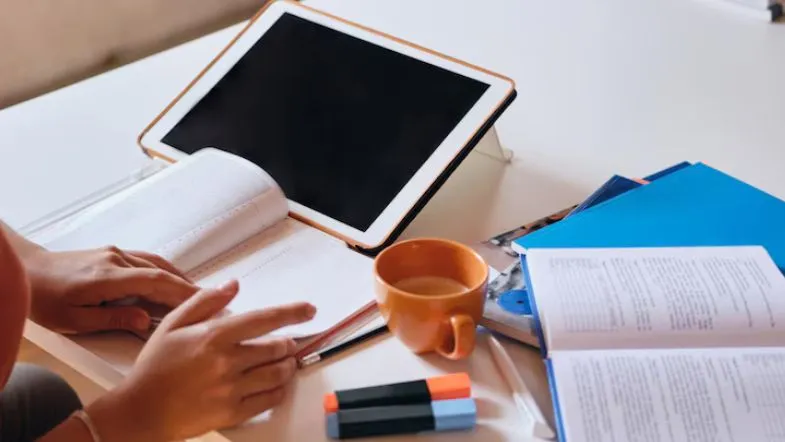 Caderno aberto e tablet em cima da mesa com canetas coloridas e uma xícara de café, representando ambiente de estudo digital.