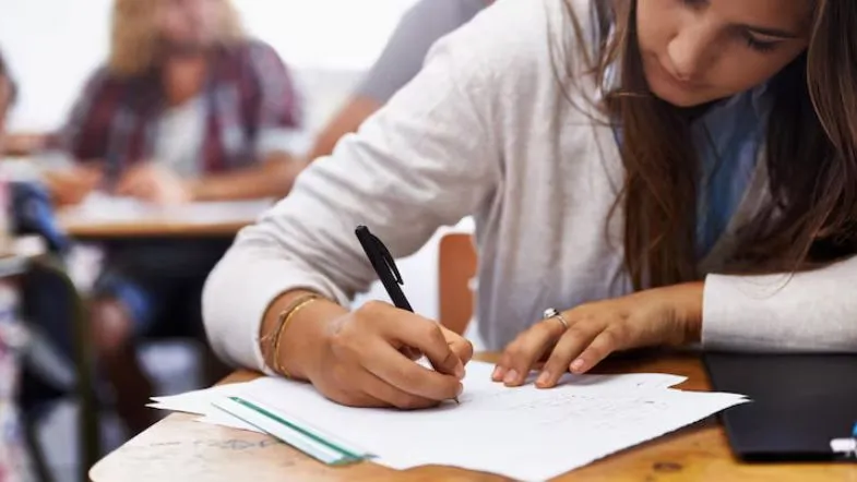 Estudante escrevendo em caderno durante prova ou atividade acadêmica em sala de aula, representando o processo seletivo.