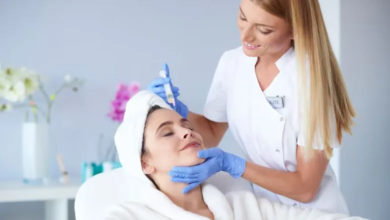 O que é Biomedicina Estética? Formação, regulamentação e áreas de atuação 13 Biomédica estética aplicando produto no rosto de paciente com touca branca e roupão, demonstrando cuidado humanizado no atendimento estético.