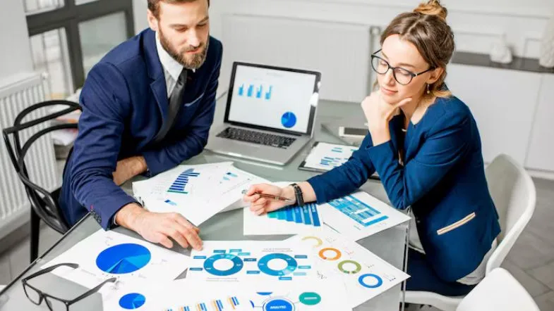Carreira em Gestão Comercial: salários, áreas de atuação e dicas para se destacar no mercado 13 Dois executivos analisando relatórios com gráficos em uma mesa de reuniões.