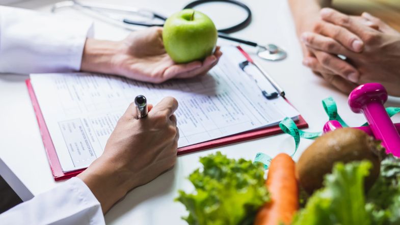 Nutricionista segurando maçã verde e preenchendo plano alimentar com alimentos naturais ao redor.