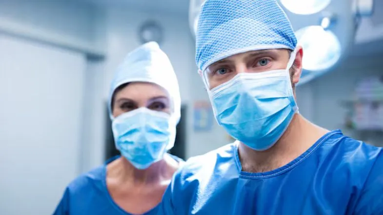 Segurança do paciente: como reduzir eventos adversos com protocolos simples 13 Cirurgiões com máscaras em ambiente hospitalar iluminado, olhando diretamente para a câmera.