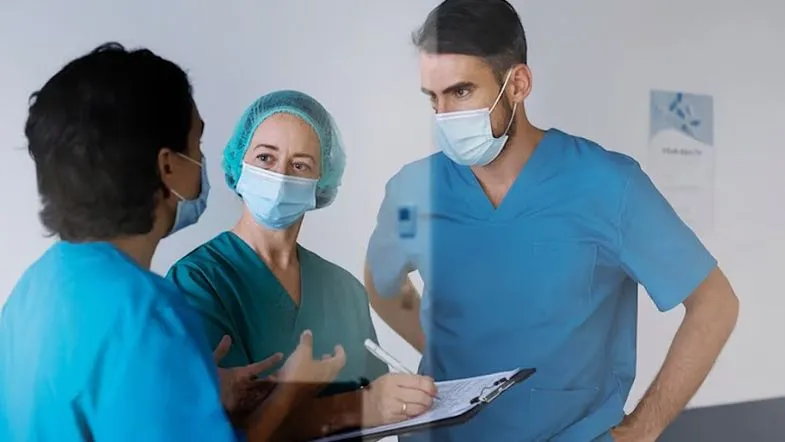 Gestão sob pressão: estratégias para formar times de alta performance 6 Equipe multiprofissional de saúde conversando com prancheta em mãos, simbolizando a gestão colaborativa em hospitais e clínicas.