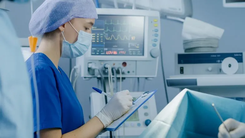 Especialização na área da saúde: conheça 6 caminhos de sucesso profissional 6 Enfermeira em unidade de terapia intensiva anotando dados de paciente em monitor hospitalar, representando a especialização em UTI.