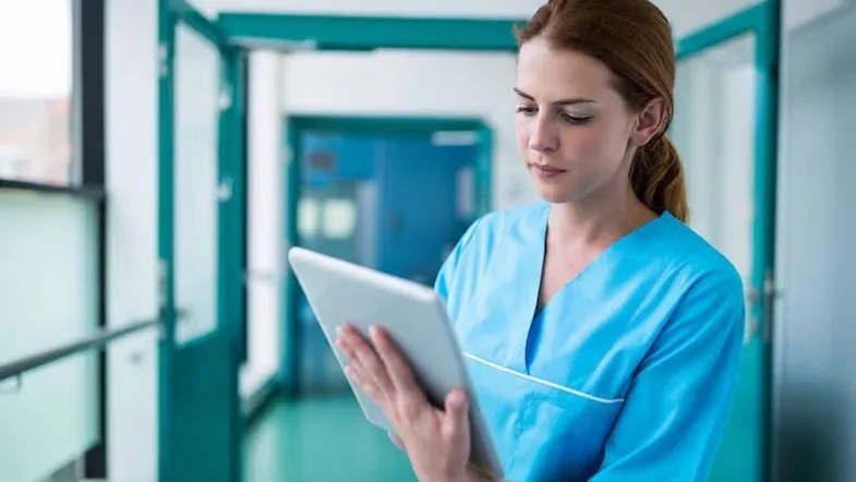Gestão sob pressão: estratégias para formar times de alta performance 7 Profissional de enfermagem utilizando tablet em corredor hospitalar, representando a aplicação da gestão digital na saúde.