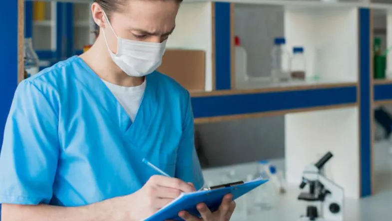 Profissional de laboratório com máscara e prancheta preenchendo registros de coleta, representando a importância da gestão da qualidade na saúde.