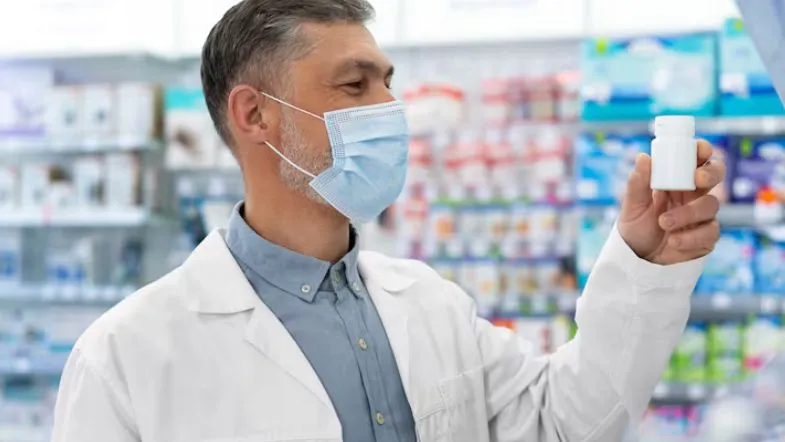 Pós-Graduação em Farmácia: veja qual área combina com o seu perfil profissional 8 Farmacêutico usando máscara e avaliando frasco de medicamento, destacando a importância da especialização em Farmácia Hospitalar.