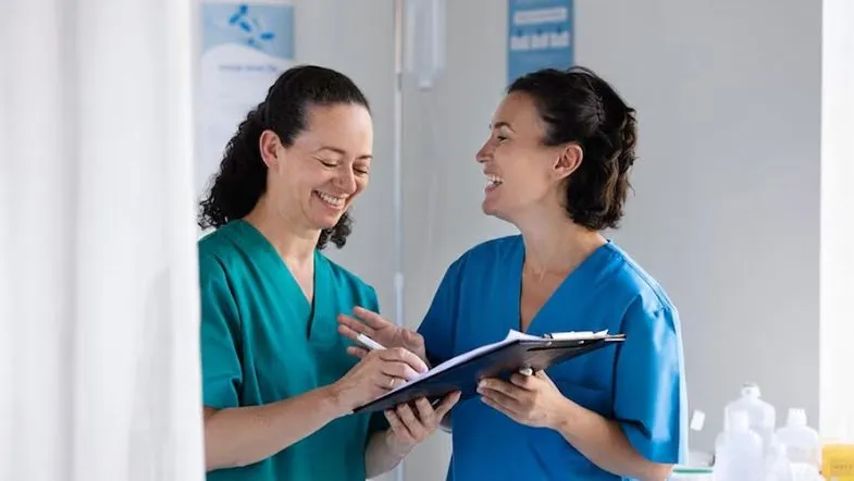 Gestão sob pressão: estratégias para formar times de alta performance 8 Duas profissionais de saúde sorrindo e revisando anotações, ilustrando a importância da gestão humanizada e do trabalho em equipe.