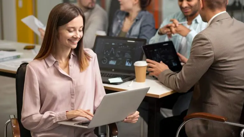 Jovem profissional de RH analisando relatórios em laptop durante reunião de equipe.