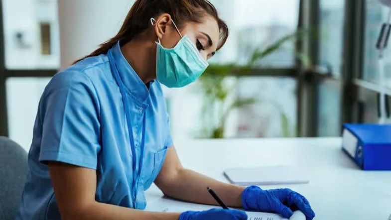 Enfermeira com máscara e luvas preenchendo prontuário durante atendimento em clínica.