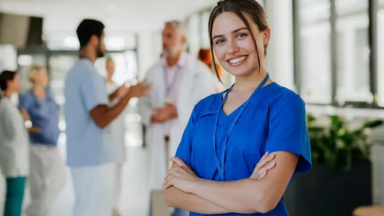 Profissional de saúde sorridente com jaleco azul em ambiente hospitalar, representando excelência em gestão, qualidade e segurança do paciente.