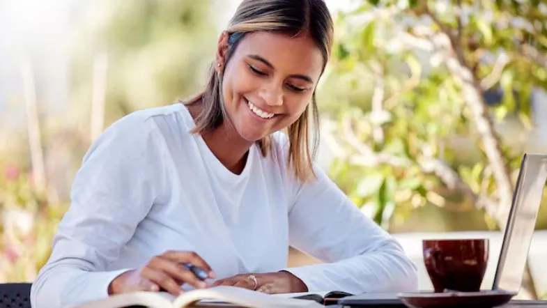 Estudante sorridente revisando conteúdos ao ar livre com livros e laptop, representando equilíbrio emocional na preparação para o ENEM 2025.