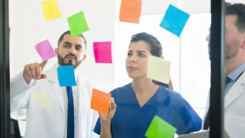 Profissionais da saúde analisando post-its coloridos em quadro transparente durante planejamento estratégico.