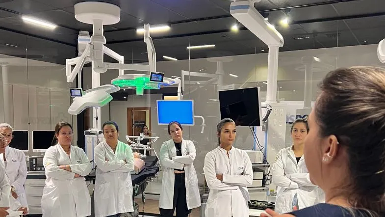 Grupo de estudantes de saúde em aula prática de Urgência e Emergência dentro de laboratório moderno, ouvindo explicação da instrutora em ambiente simulado de urgência.