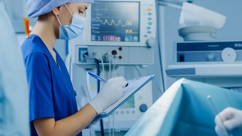 Carreira na UTI Neonatal em 2025: remuneração, oportunidades e o papel da pós-graduação 7 Profissional de saúde com máscara preenchendo prontuário em sala de UTI equipada com monitor multiparamétrico.