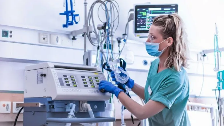 Apgar, reanimação e ventilação: o guia completo da prática em UTI Neonatal e Pediátrica em 2025 8 Enfermeira com máscara ajustando ventilador mecânico em ambiente de UTI.