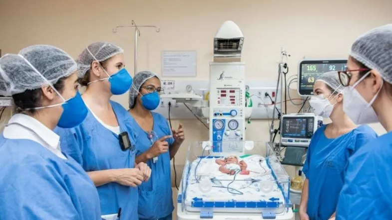 Quanto dura a pós em UTI Neonatal e Pediátrica? Entenda a duração ideal 9 Equipe multiprofissional reunida ao redor de incubadora com recém-nascido em UTI Neonatal, usando máscaras e EPIs.
