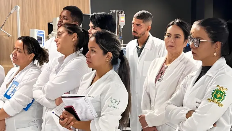 Grupo de alunos vestindo jalecos observando explicação em laboratório com equipamentos de simulação e dispositivos hospitalares.