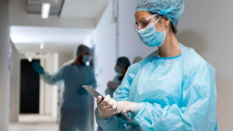 Profissional de saúde com EPIs utilizando tablet em corredor hospitalar, integrando dados clínicos.