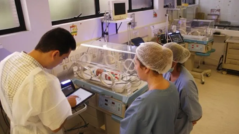 Equipe de profissionais de saúde avaliando um recém-nascido em incubadora dentro da UTI Neonatal.