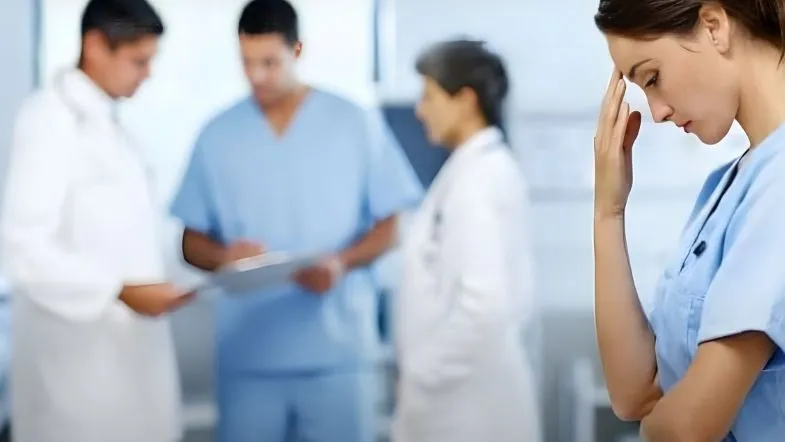 Profissional de enfermagem preocupada, com equipe médica desfocada ao fundo, ilustrando desgaste e esgotamento em serviços de saúde.