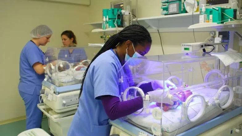 Enfermeira atuando em UTI Neonatal, realizando monitorização e cuidados intensivos em incubadora neonatal.