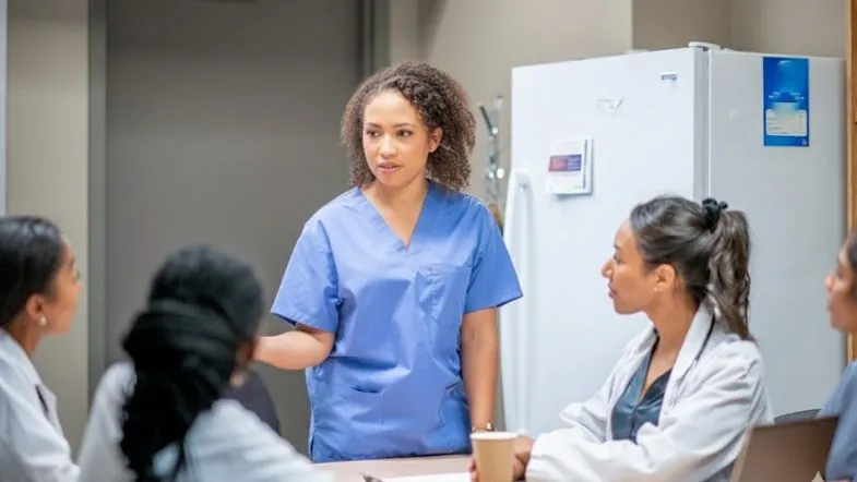 Profissional em enfermagem liderando reunião clínica com equipe multiprofissional, representando comunicação e gestão humanizada.