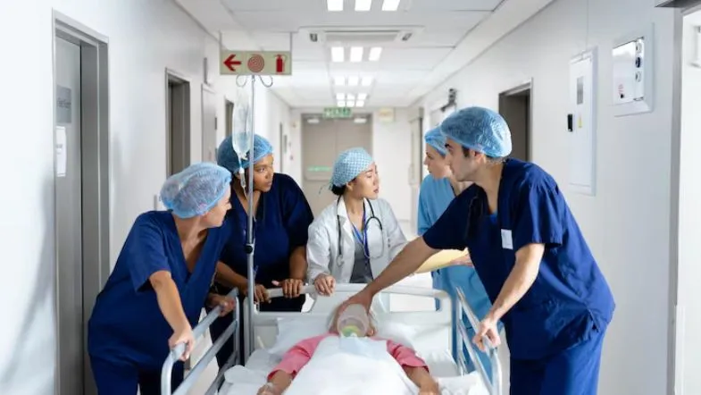 Equipe multiprofissional transportando paciente em maca por corredor hospitalar durante atendimento de Urgência e Emergência.