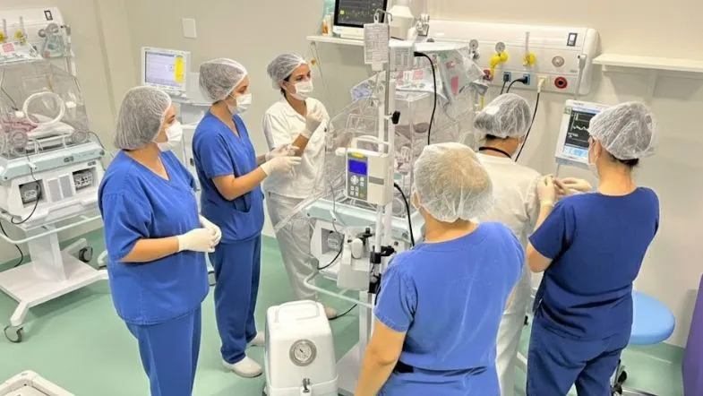 Grupo de profissionais reunidos em torno de incubadora em UTI Neonatal participando de atividade de capacitação e monitorização clínica.