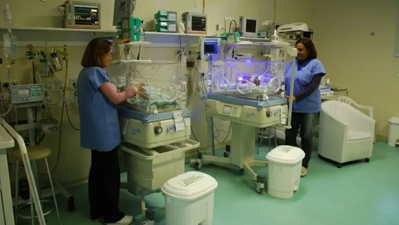 Equipe de enfermagem atuando em UTI Neonatal com múltiplas incubadoras e equipamentos de suporte intensivo ao prematuro.
