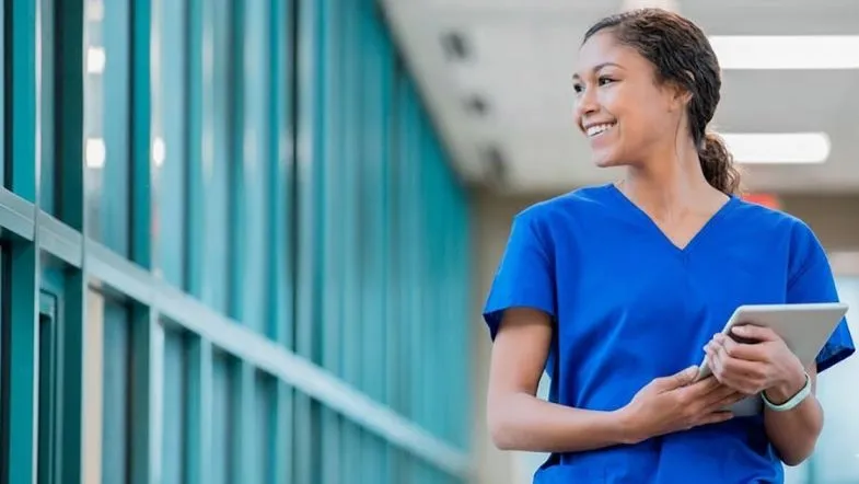 Profissional da saúde sorrindo, com tablet em ambiente hospitalar, representando desenvolvimento profissional e liderança estratégica.