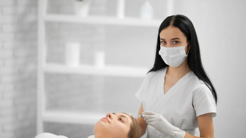 Profissional com máscara aplicando injetável na região facial de paciente durante tratamento estético.
