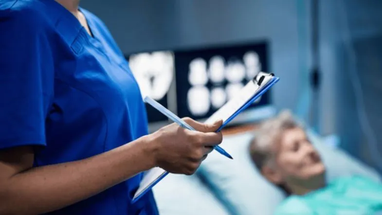 Qualidade e segurança na enfermagem: por que a gestão estratégica é a prioridade em 2026 6 Close-up das mãos de uma enfermeira segurando uma prancheta e caneta. Ao fundo, uma tela de monitor exibe exames de imagem e gráficos assistenciais.