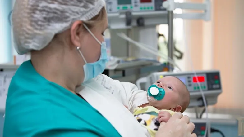 Profissional de saúde com máscara e touca segurando um bebê recém-nascido com chupeta em ambiente de unidade de terapia intensiva neonatal.