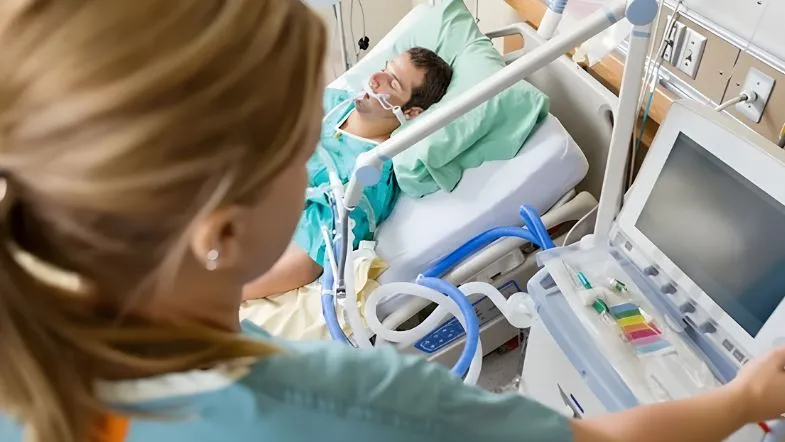Visão por cima do ombro de uma enfermeira loira observando um paciente masculino sob ventilação mecânica invasiva. Ela estende a mão para ajustar o monitor do ventilador mecânico ao lado do leito, representando o ambiente da UTI.