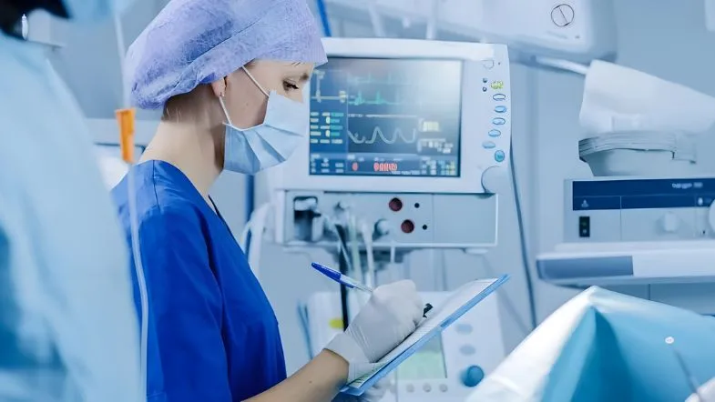 Enfermeira de perfil usando touca e máscara azul claro, escrevendo atentamente em um documento hospitalar. Ao fundo, telas de monitores multiparamétricos exibem curvas de sinais vitais de forma nítida.