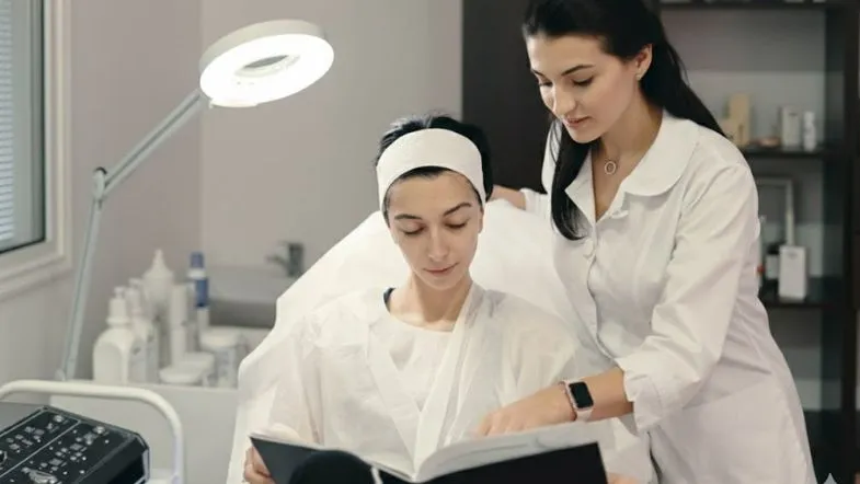 Especialista em estética segurando uma prancheta e analisando a pele de uma paciente deitada. Ao fundo, monitores de equipamentos de estética avançada.
