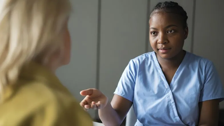Qualidade e segurança na enfermagem: por que a gestão estratégica é a prioridade em 2026 7 Uma enfermeira com uniforme azul conversa de forma acolhedora e atenta com uma paciente, utilizando gestos manuais para explicar um procedimento.