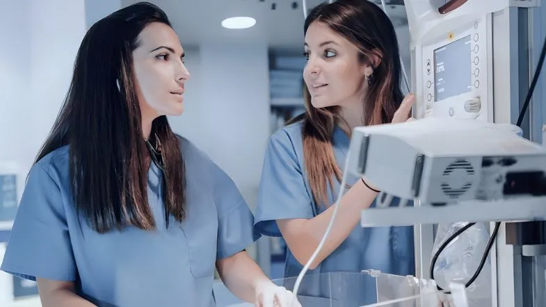 Duas enfermeiras especialistas discutindo dados clínicos em monitor multiparamétrico de alta tecnologia em uma UTI Neonatal e Pediátrica moderna.