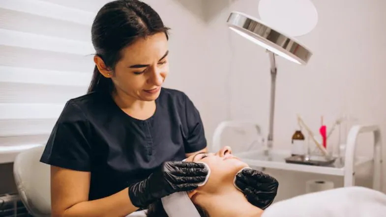 Estética Avançada em Goiânia e Brasília: onde se especializar em 2026 8 Profissional com luvas pretas realizando limpeza e preparação da pele de uma paciente sob luz de foco clínico.