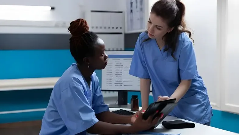 Qualidade e segurança na enfermagem: por que a gestão estratégica é a prioridade em 2026 8 Duas enfermeiras em um posto de enfermagem moderno. Uma delas segura um tablet e aponta para a tela, enquanto a outra observa atentamente. Ao fundo, um monitor de computador com dados de gestão.