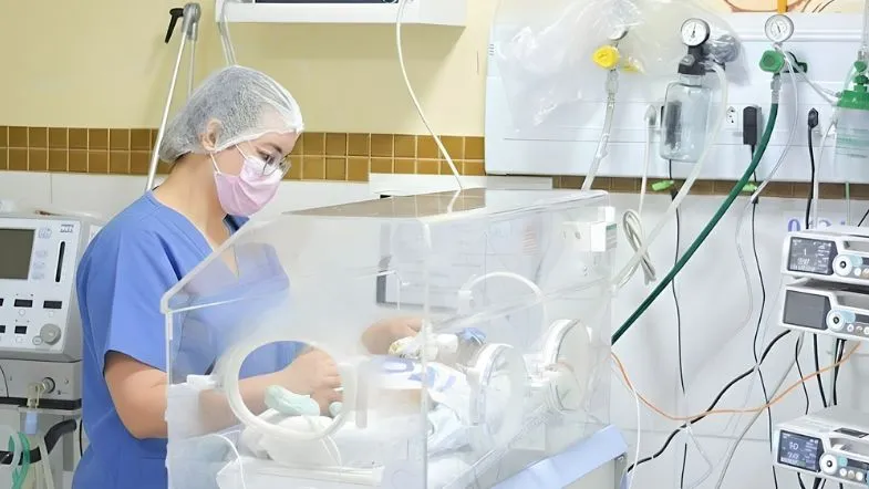 Enfermeira intensivista realizando manuseio técnico em incubadora de UTI neonatal de alta complexidade com equipamentos de monitoramento ao fundo.