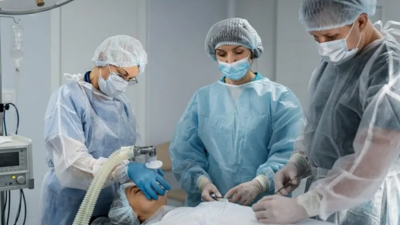 Equipe médica composta por três profissionais de UTI paramentados com capotes azuis e cinzas, máscaras e toucas, realizando um procedimento de intubação ou manejo de via aérea em paciente sedado.