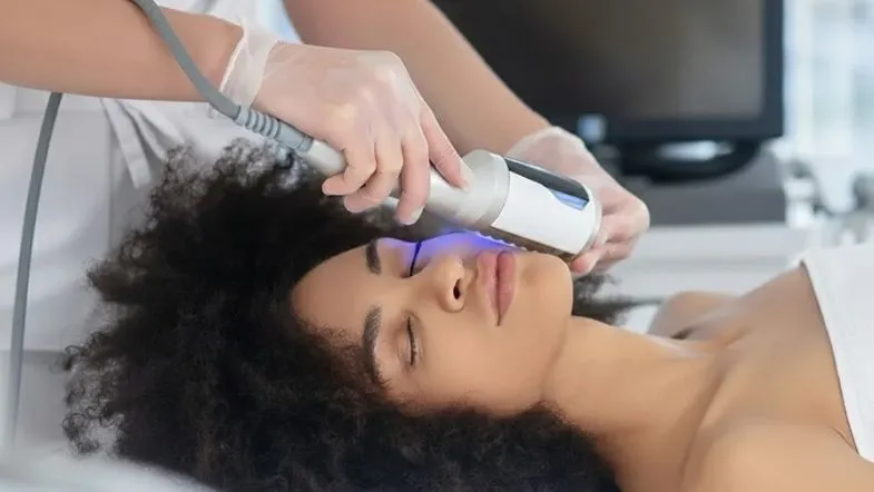 Especialista realizando procedimento tecnológico na face de uma paciente jovem de pele negra e cabelo afro. Foco na precisão do aplicador próximo ao olho.