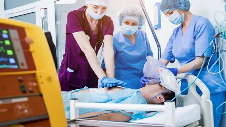 Urgência, Emergência e UTI: como a prática clínica diferencia o enfermeiro especialista 9 Profissionais de saúde realizando ventilação com ambu e massagem cardíaca em um paciente, com um desfibrilador em destaque no primeiro plano.