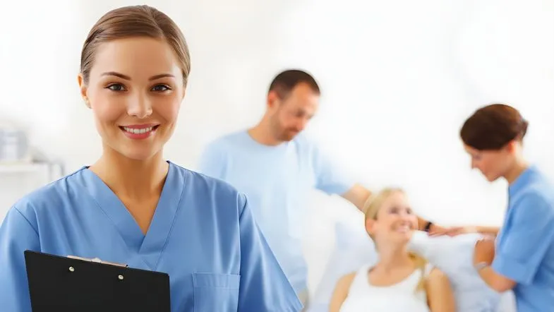 Qualidade e segurança na enfermagem: por que a gestão estratégica é a prioridade em 2026 9 Uma enfermeira sorridente em primeiro plano segurando uma pasta preta. Ao fundo, de forma desfocada, outros profissionais de saúde realizam o atendimento de uma paciente em um leito.